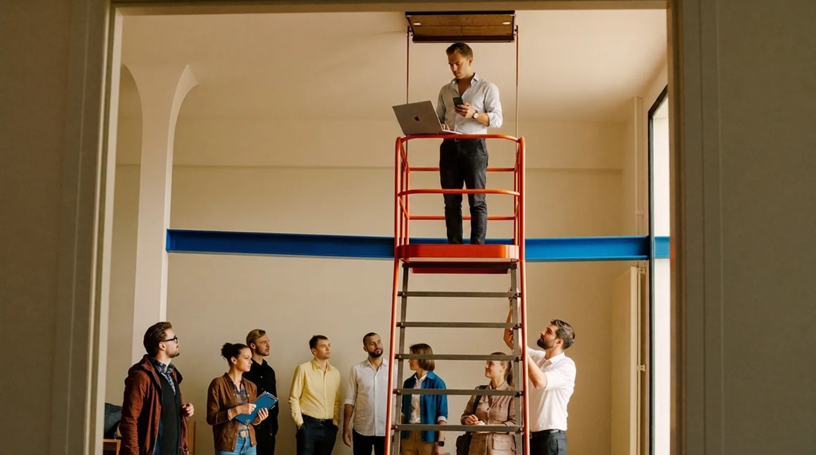Een ondernemer staat hoog op een hoogwerker druk te werken met laptop en telefoon, terwijl zijn team beneden op hem staat te wachten